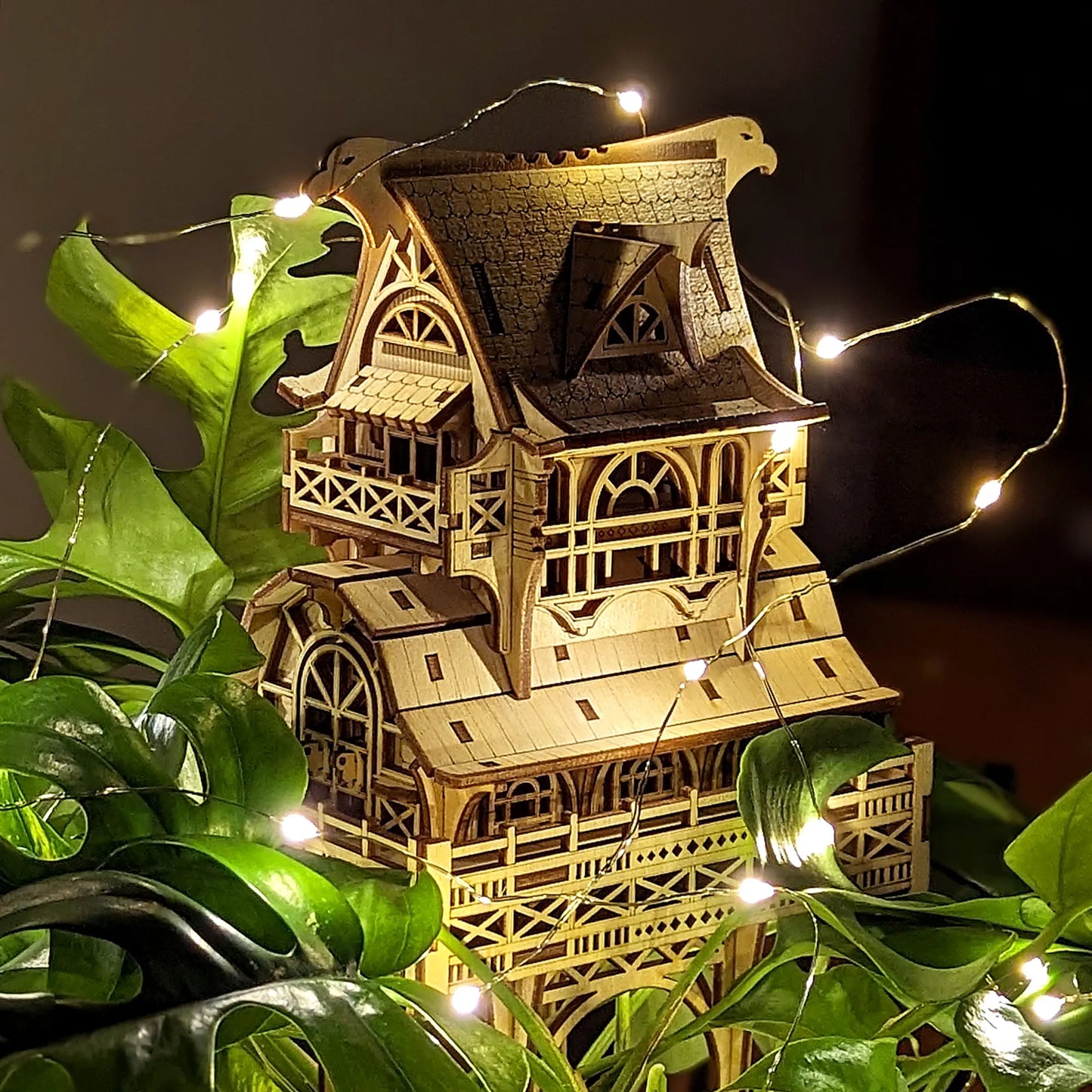 Shaded side view of a laser cut plywood fantasy house model placed among dark green leaves with glowing mini lights.