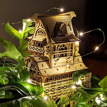 Shaded side view of a laser cut plywood fantasy house model placed among dark green leaves with glowing mini lights.