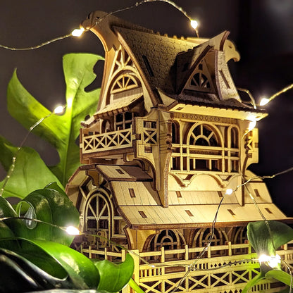 Close up of a laser cut plywood fantasy house model glowing under warm fairy lights next to indoor green leaves.