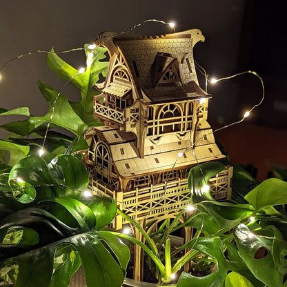 Detailed laser cut plywood miniature house nestled inside a large green houseplant and decorated with LED lights.