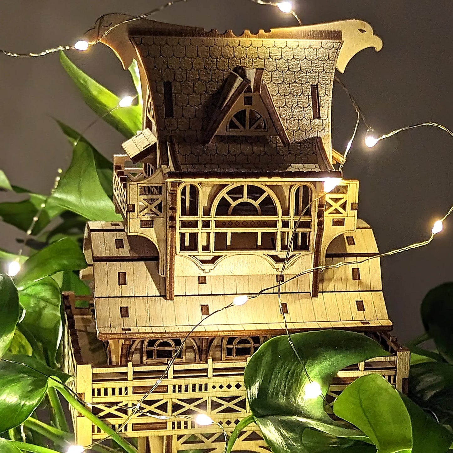 Center view of a laser cut plywood miniature house featuring an engraved shingle roof, balconies, and fairy lights.