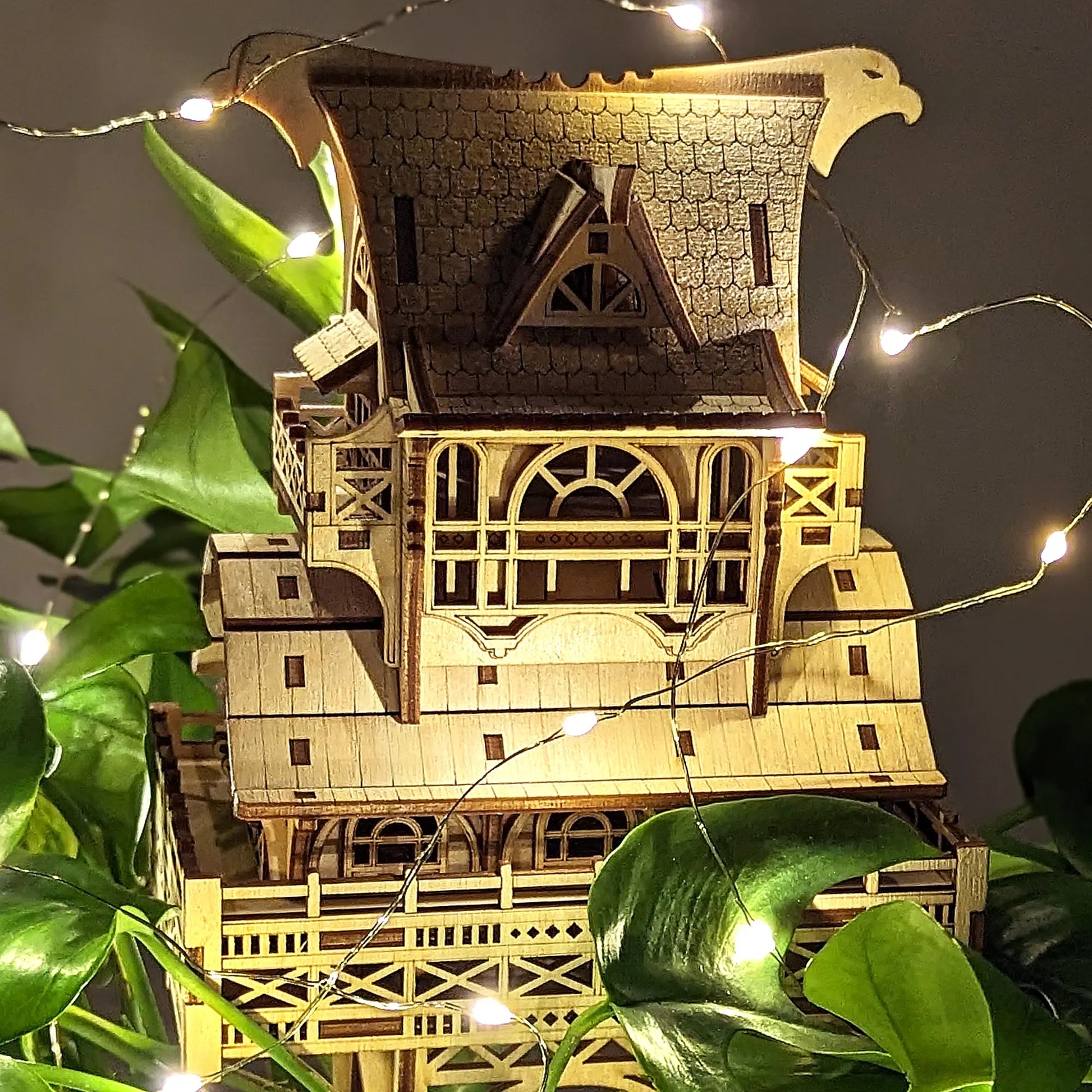 Center view of a laser cut plywood miniature house featuring an engraved shingle roof, balconies, and fairy lights.