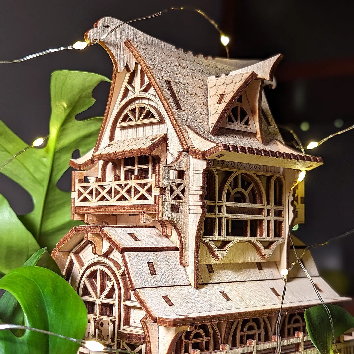 Three-quarter view of a laser cut plywood fantasy cottage wrapped in warm fairy lights next to a monstera plant.