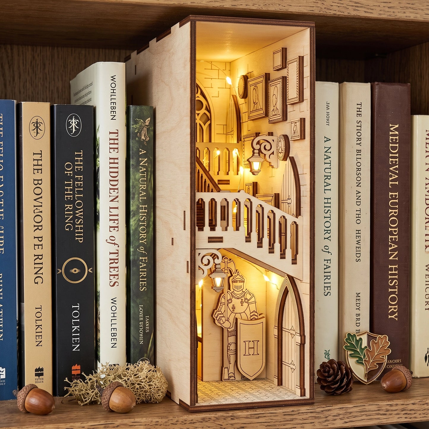 Lit laser-cut birch plywood castle book nook on a shelf with fantasy books, acorns, and moss, showing warm interior lights.