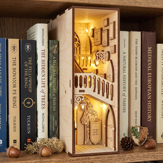 Lit laser-cut birch plywood castle book nook on a shelf with fantasy books, acorns, and moss, showing warm interior lights.