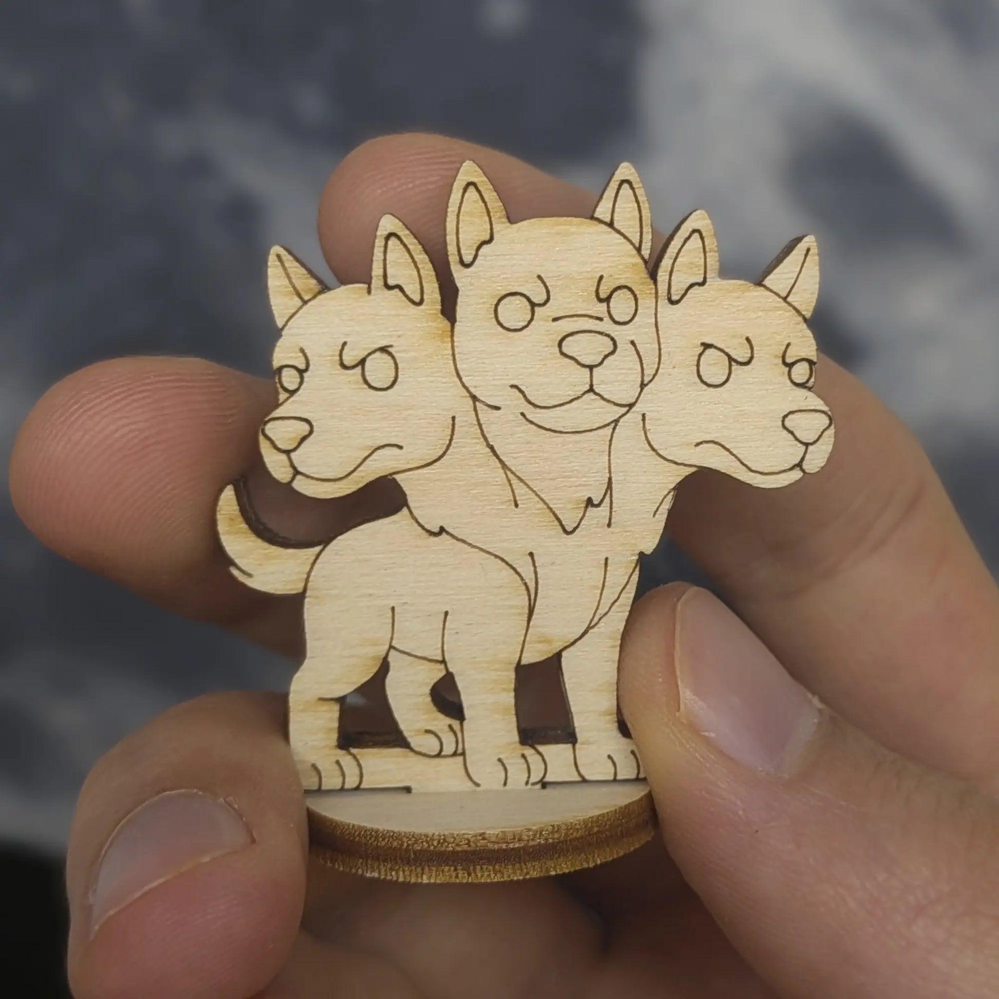 Hand holds a 3D Cerberus model of laser cut plywood. Front view shows three heads, paws, and detailed engraved features