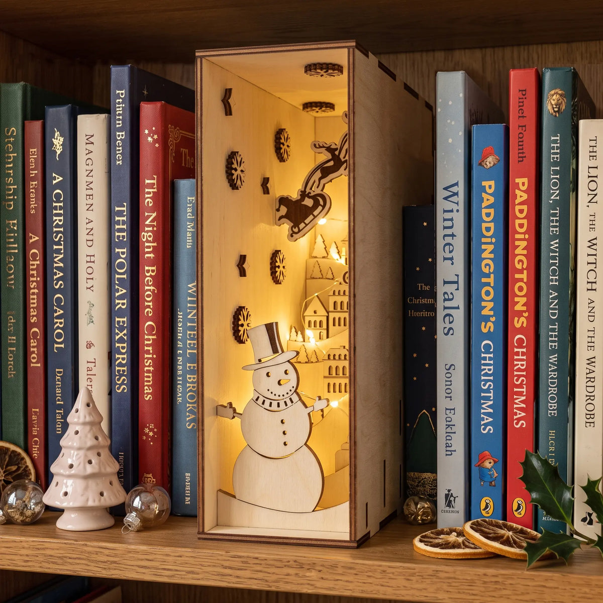 Lit laser cut plywood holiday book nook diorama displayed between books on wooden shelf with festive decor.