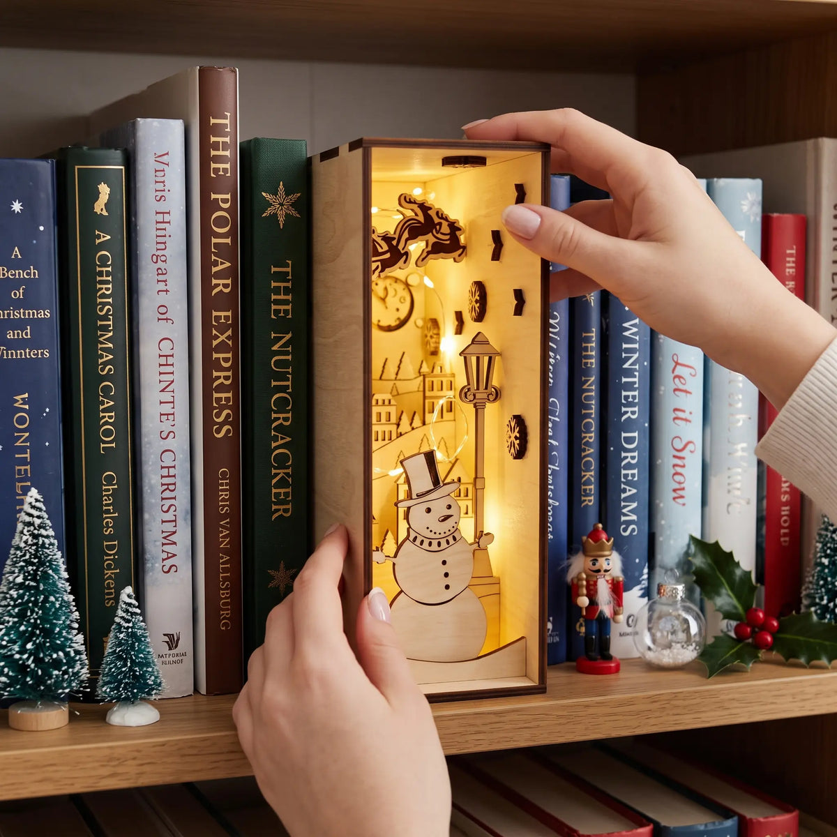 Hand places glowing laser cut plywood holiday book nook diorama with snowman and Santa sleigh into bookshelf