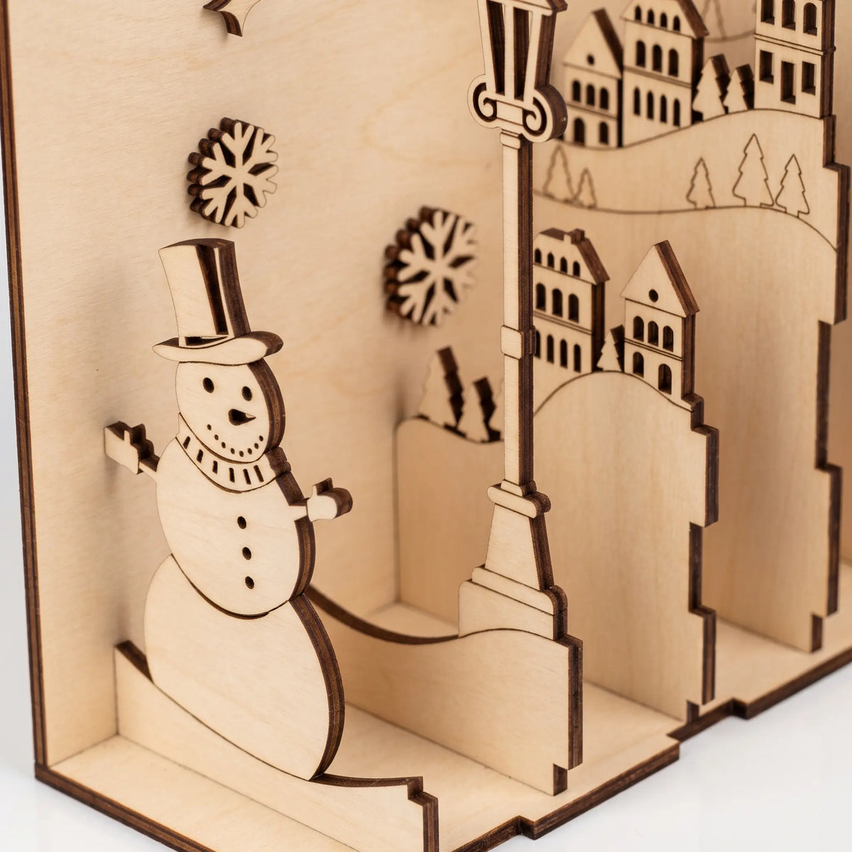 Close-up of layered laser cut plywood book nook diorama showing slotted snowman, lamppost, and hillside village panels.