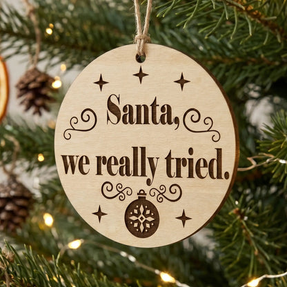 Round laser cut plywood Christmas ornament with 'Santa, we really tried' quote, hanging on a pine branch with warm lights.
