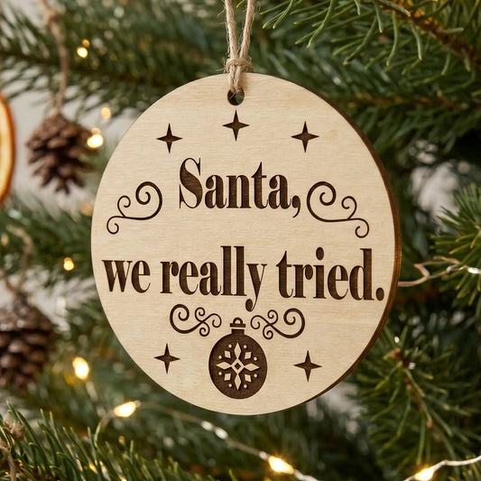 Round laser cut plywood Christmas ornament with 'Santa, we really tried' quote, hanging on a pine branch with warm lights.