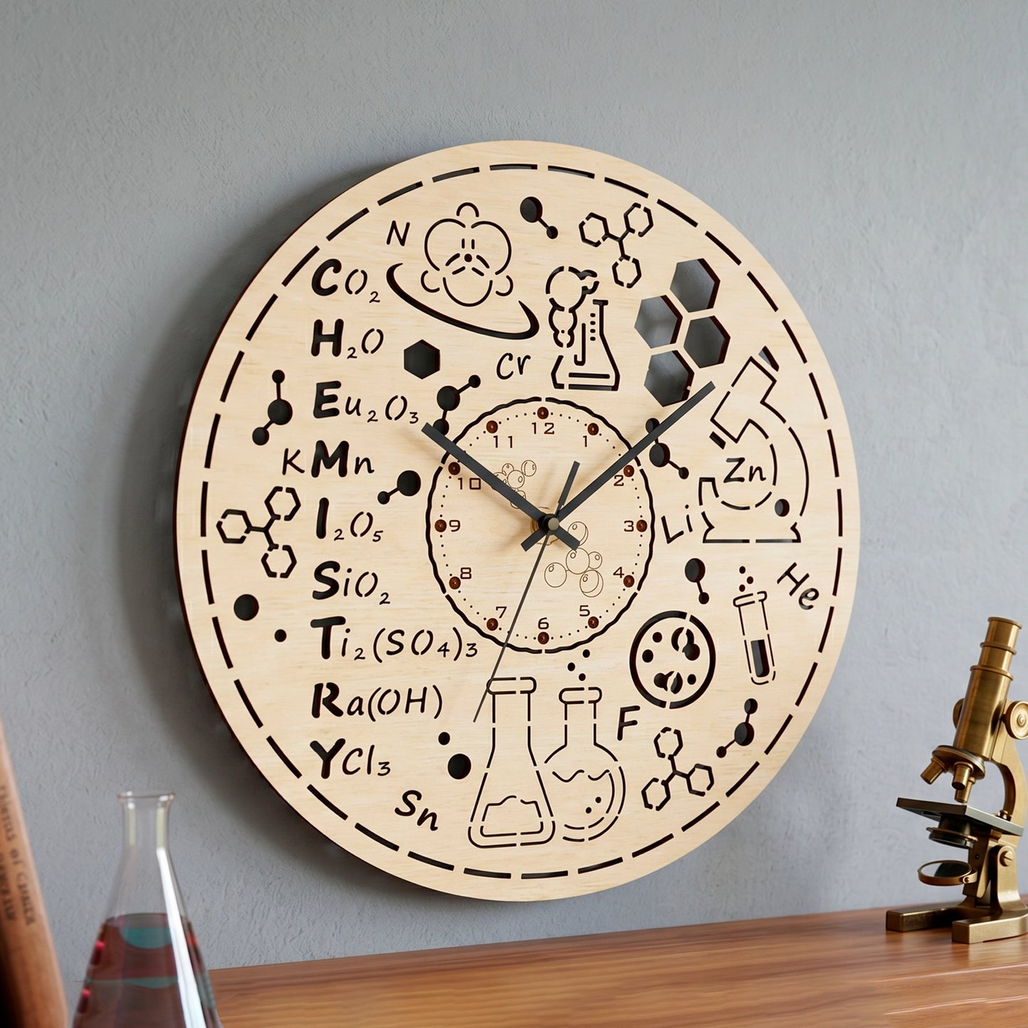 Laser cut plywood science clock on a gray wall above a wooden desk with a vintage brass microscope and glass flask.