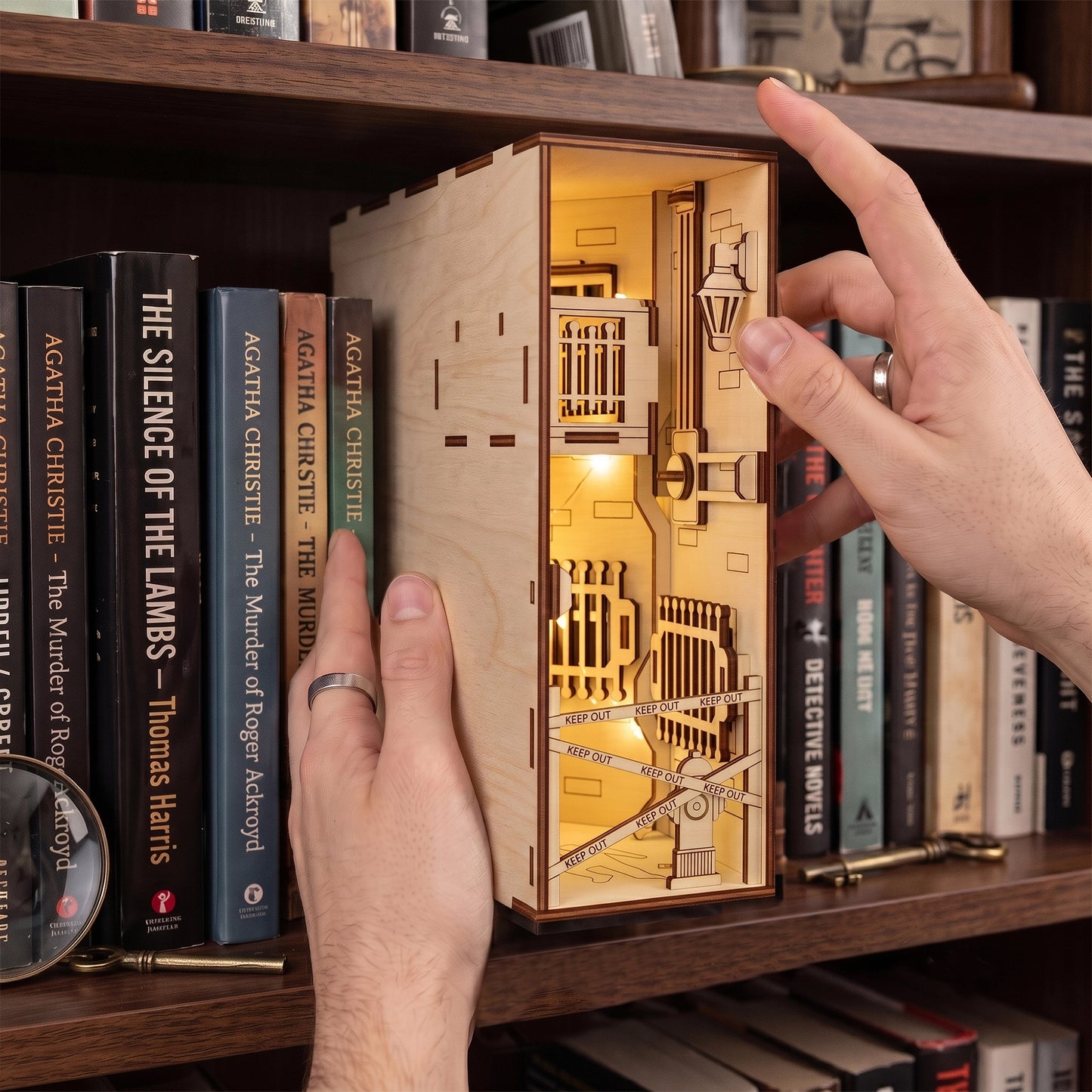 Hands placing a lit laser-cut birch plywood crime scene book nook on a shelf next to detective novels and props.