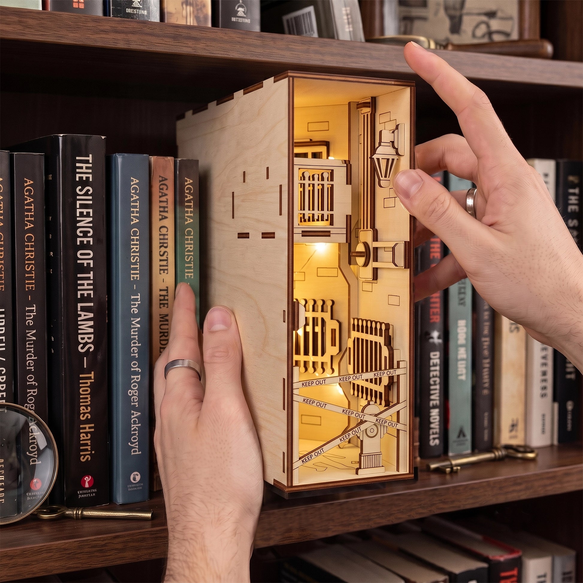 Hands placing a lit laser-cut birch plywood crime scene book nook on a shelf next to detective novels and props.