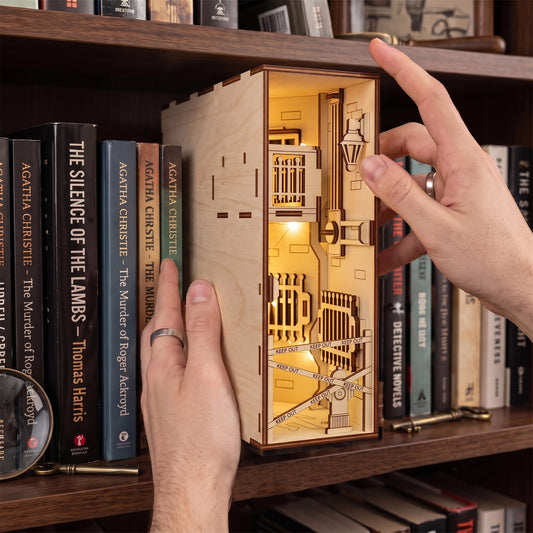Hands placing a lit laser-cut birch plywood crime scene book nook on a shelf next to detective novels and props.