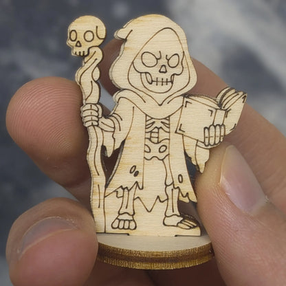 Hand holds a laser cut plywood dark necromancer. Shows a skull staff, tattered robes, and skeletal face for tabletop scale