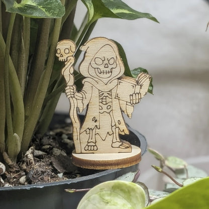 A dark necromancer of laser cut plywood stands in a plant pot. Features a skull staff and tattered robes on natural wood