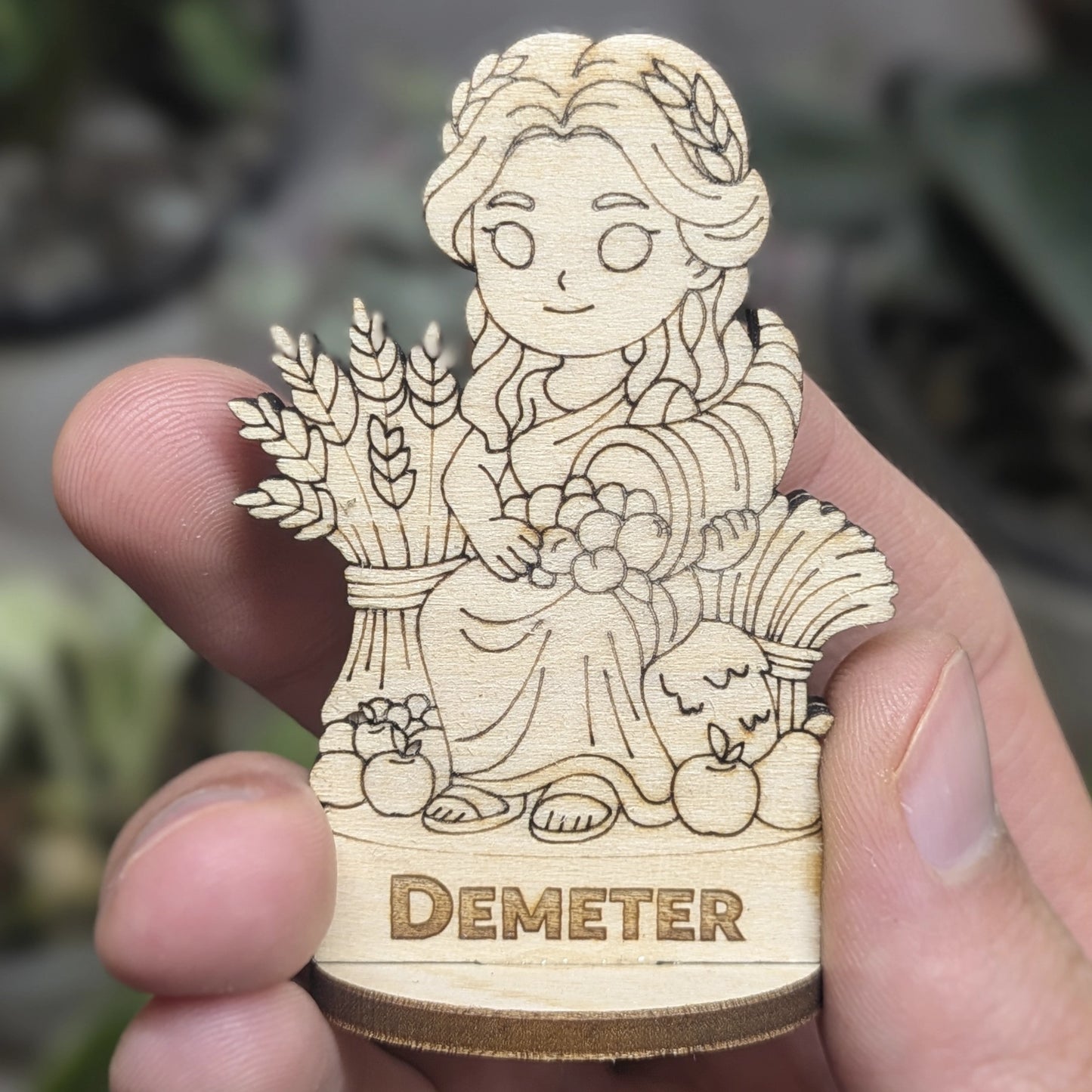 Hand holds a laser cut plywood Demeter for scale. Detailed front shows the engraved sheaf of grain, crown, and flowing robes