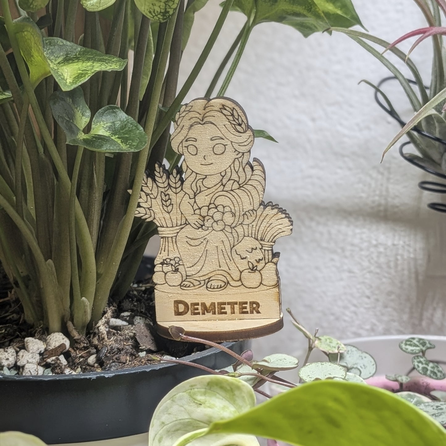 Laser cut plywood Demeter figurine sits in a plant pot. Features engraved wheat sheaf, flowing robes, and natural wood grain