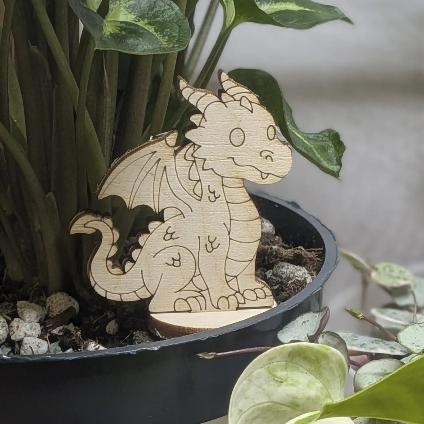 Laser cut plywood dragon figurine staged in a plant pot. Detailed engravings of wings and scales stand out against leaves