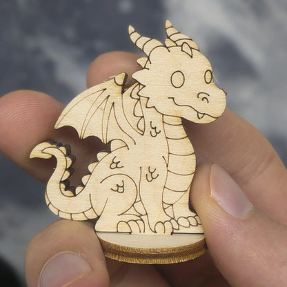 Hand holds a small dragon of laser cut plywood. Features engraved scales, wings, horns, and a cute face on a round wood base