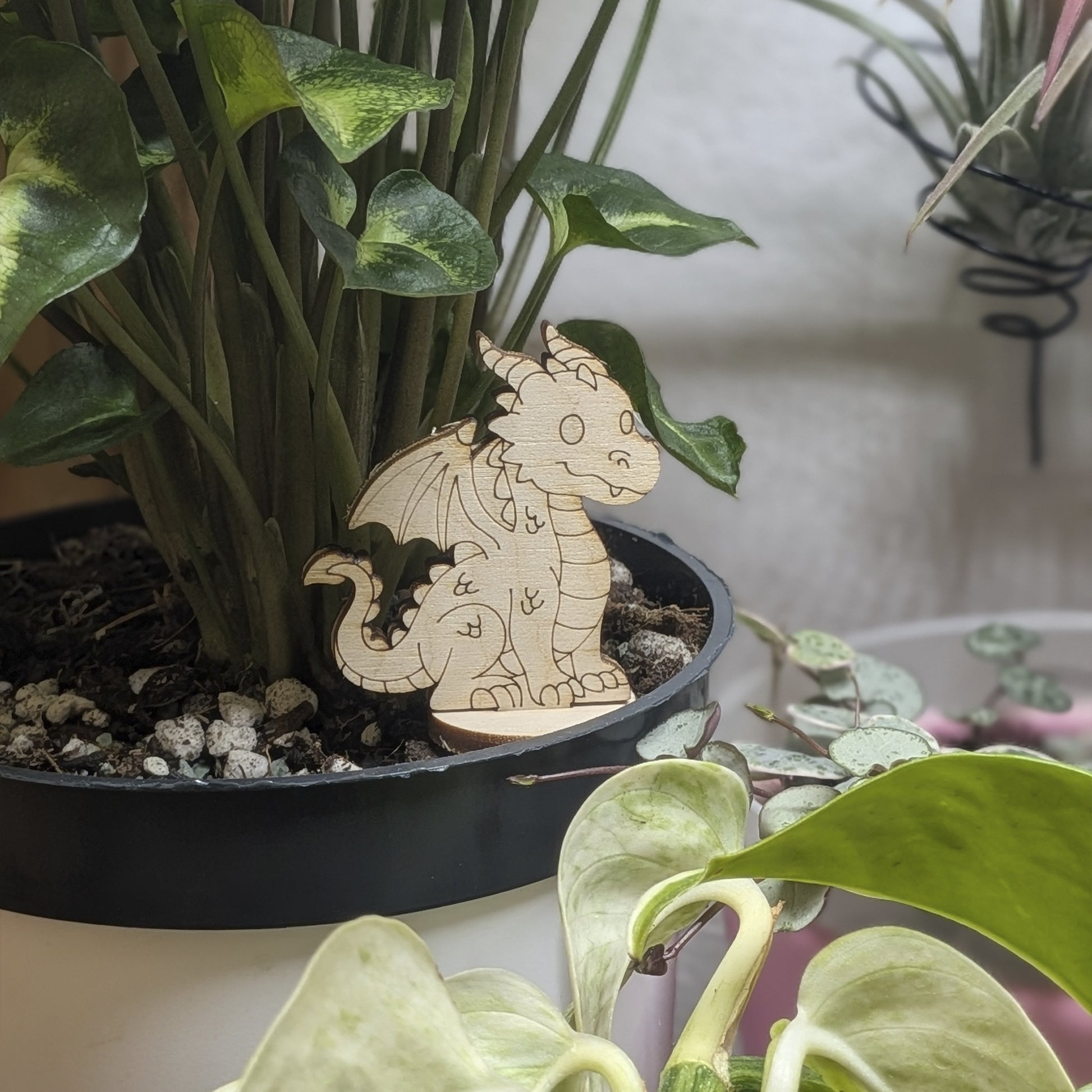 Laser cut plywood dragon figurine staged in a plant pot. Detailed engravings of wings and scales stand out against leaves