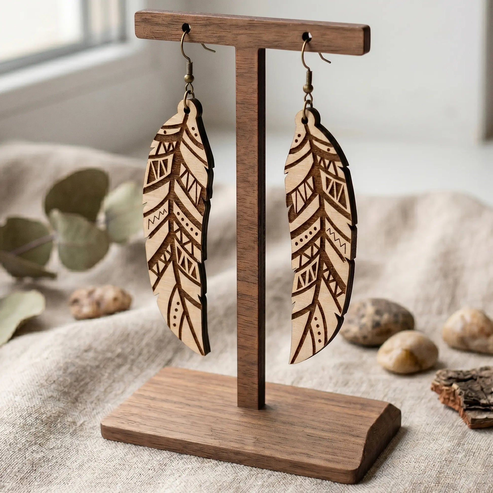 Boho laser cut plywood feather earrings with geometric engravings hanging on a wooden stand over rustic linen and stones
