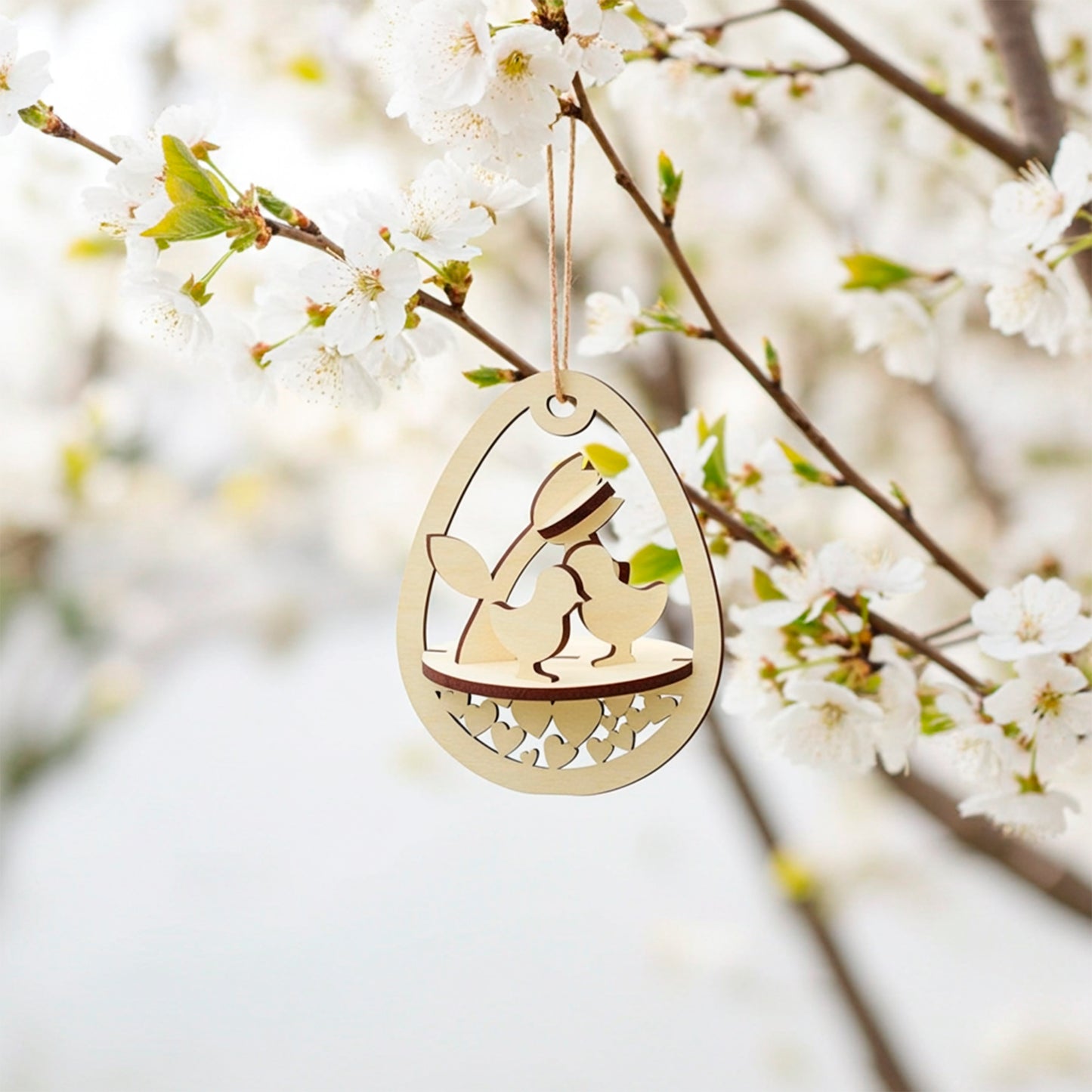 Laser-cut plywood wooden Easter egg ornament hanging from a white flowering cherry branch. The egg-shaped frame features a layered scene inside with two baby chicks, a tulip, and a base decorated with cutout hearts