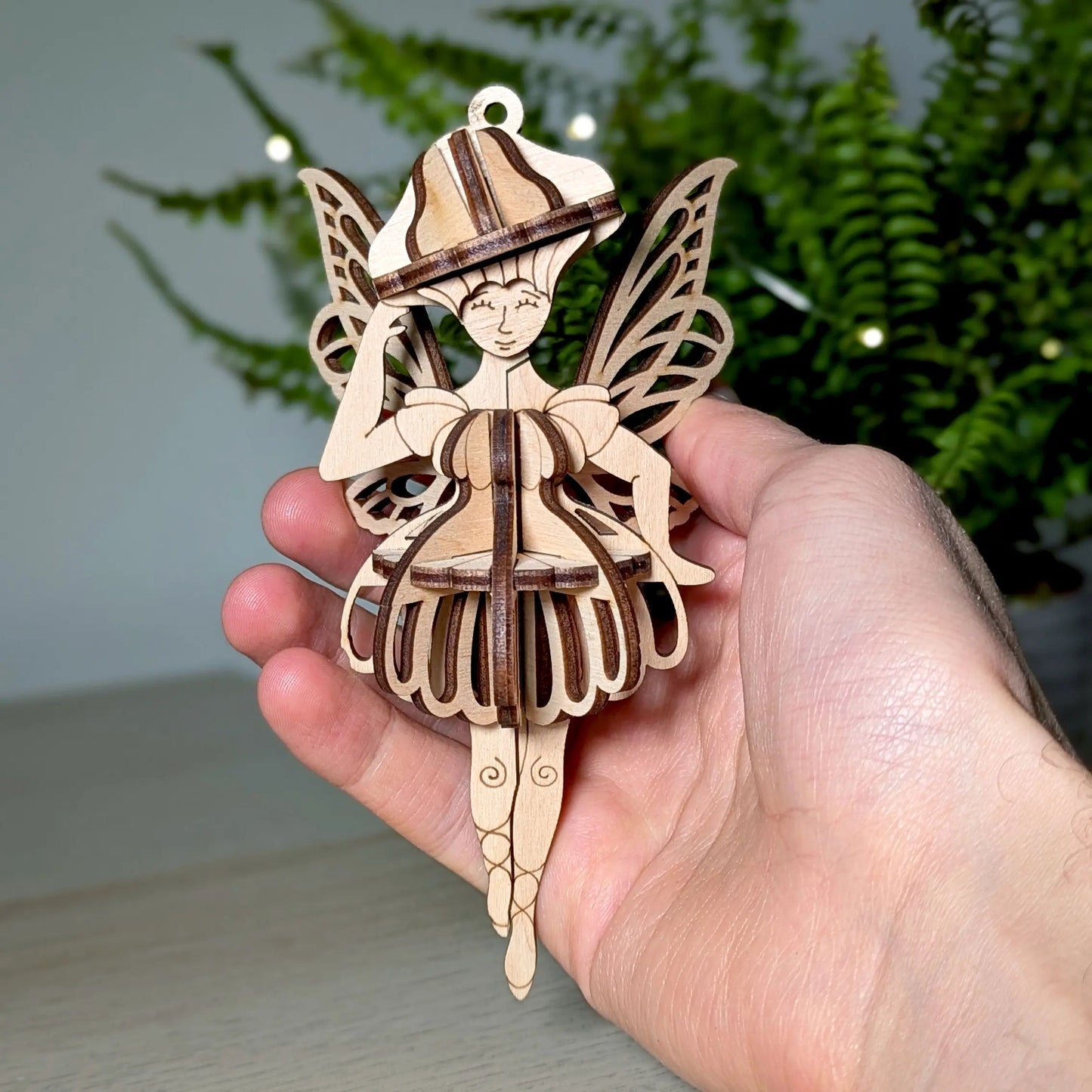 A 3D laser cut plywood fairy ornament held in a hand to show scale, with a green plant background.