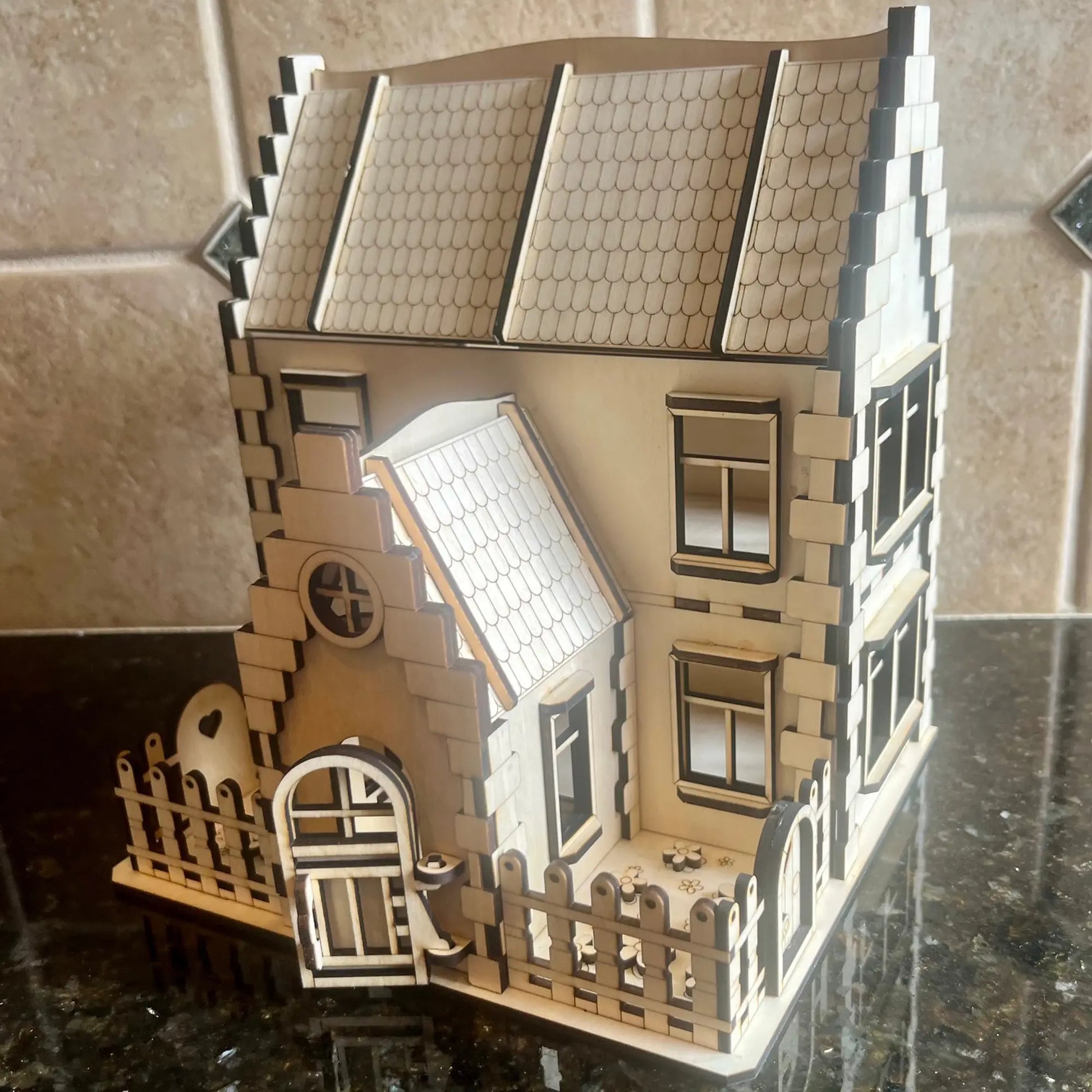 Front angle view of an assembled laser cut plywood miniature family house featuring an open porch door and a fenced yard.