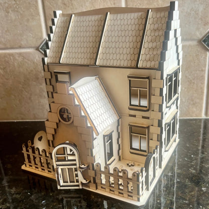 Front angle view of an assembled laser cut plywood miniature family house featuring an open porch door and a fenced yard.