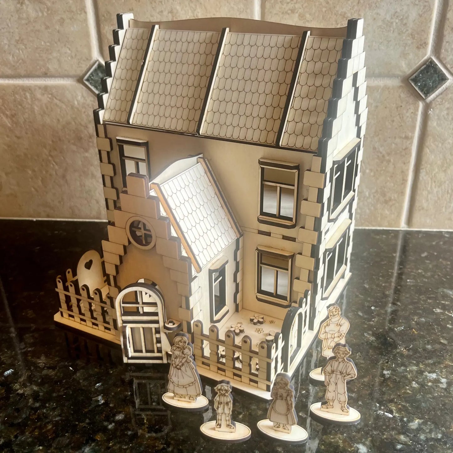 Laser cut plywood family house displayed on a dark counter with five miniature wooden family figurines standing out front.