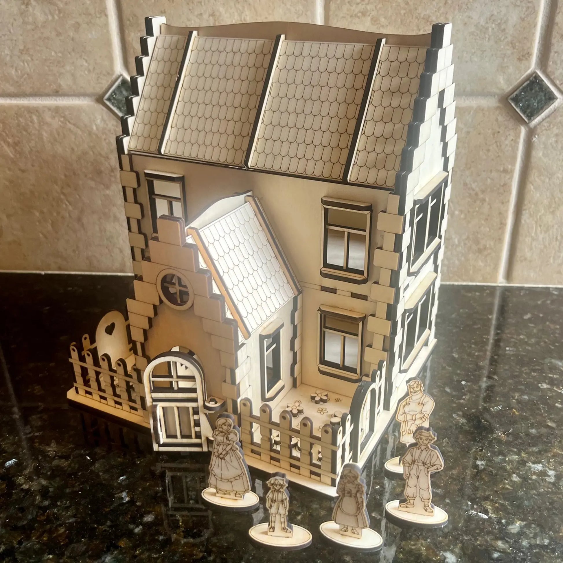 Laser cut plywood family house displayed on a dark counter with five miniature wooden family figurines standing out front.