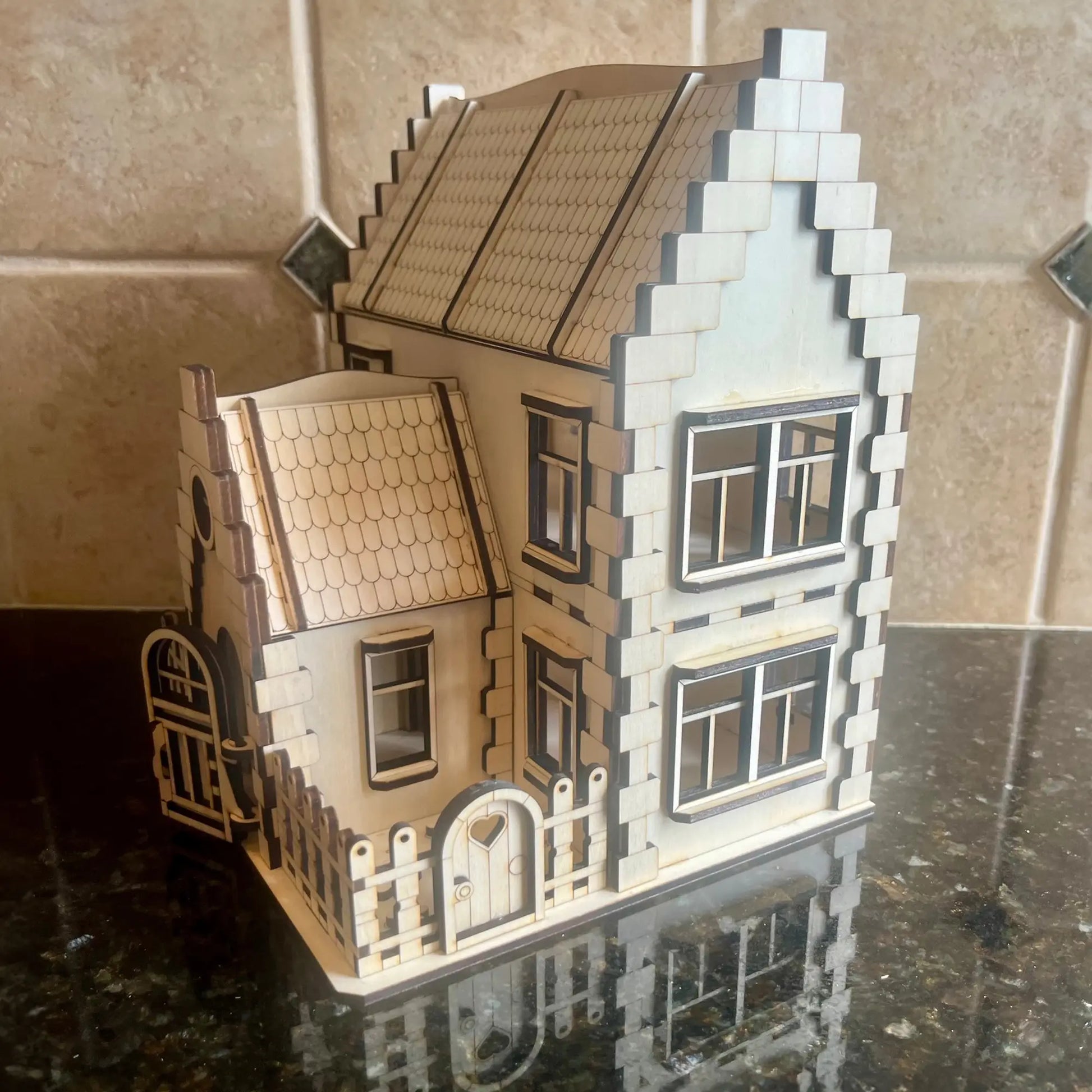 Assembled laser cut plywood family dollhouse with a small fenced yard and heart gate, displayed on a glossy dark surface.