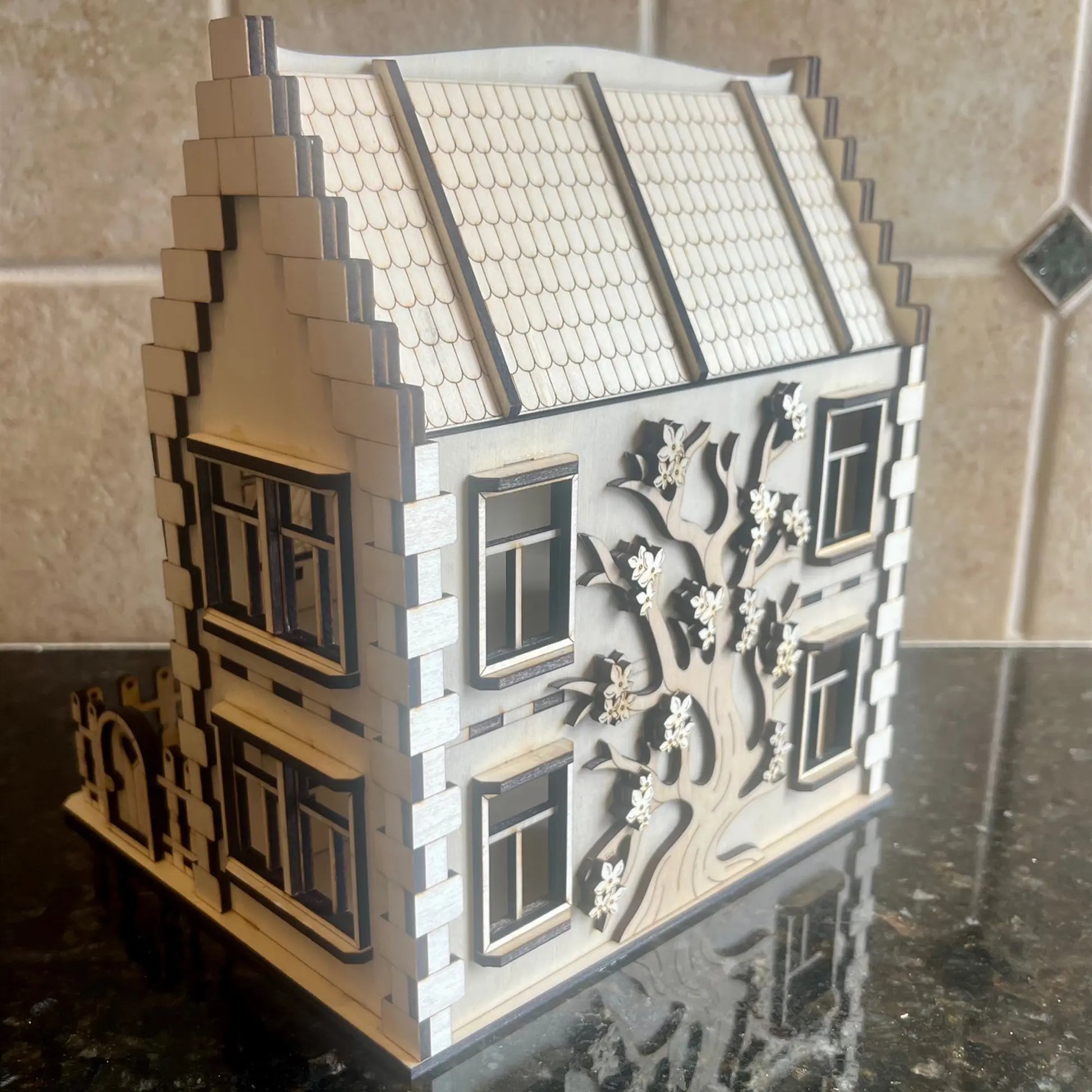 Side view of a laser cut plywood miniature house showcasing an engraved blooming tree on the wall, on a dark countertop.