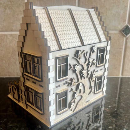 Side view of a laser cut plywood miniature house showcasing an engraved blooming tree on the wall, on a dark countertop.