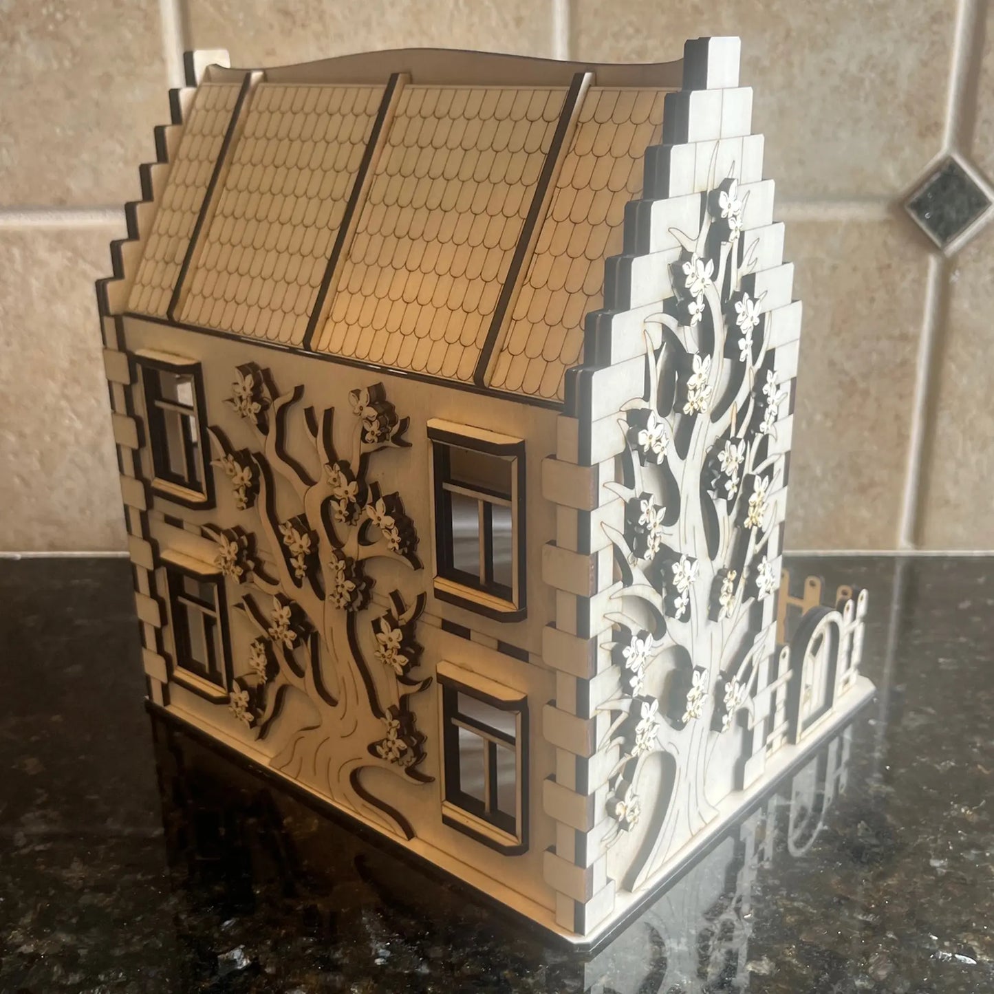 Back angle view of a laser cut plywood family house featuring a detailed engraved tree design across the entire wooden wall.