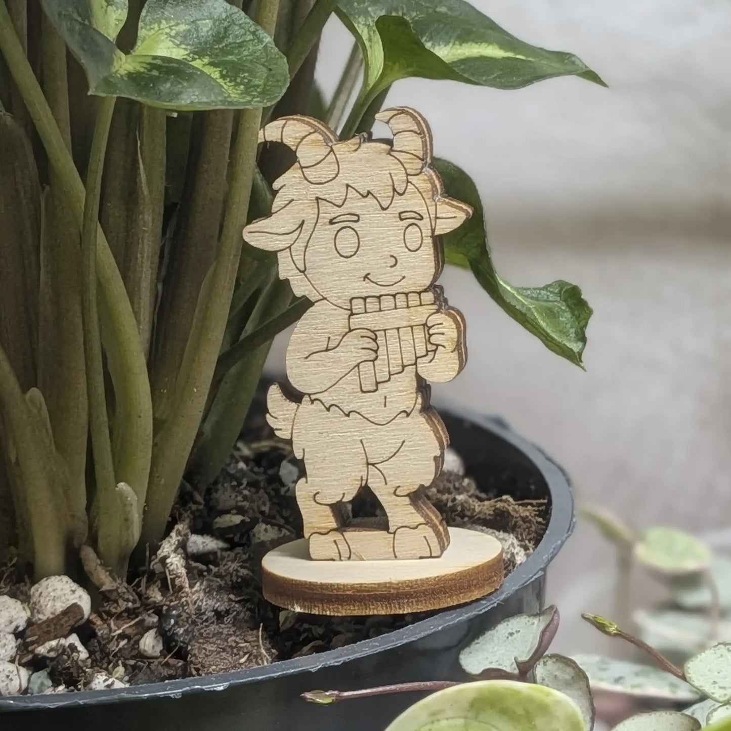 Laser cut plywood faun figurine staged in a plant pot. He plays panpipes amidst green leaves on a sturdy round wood base