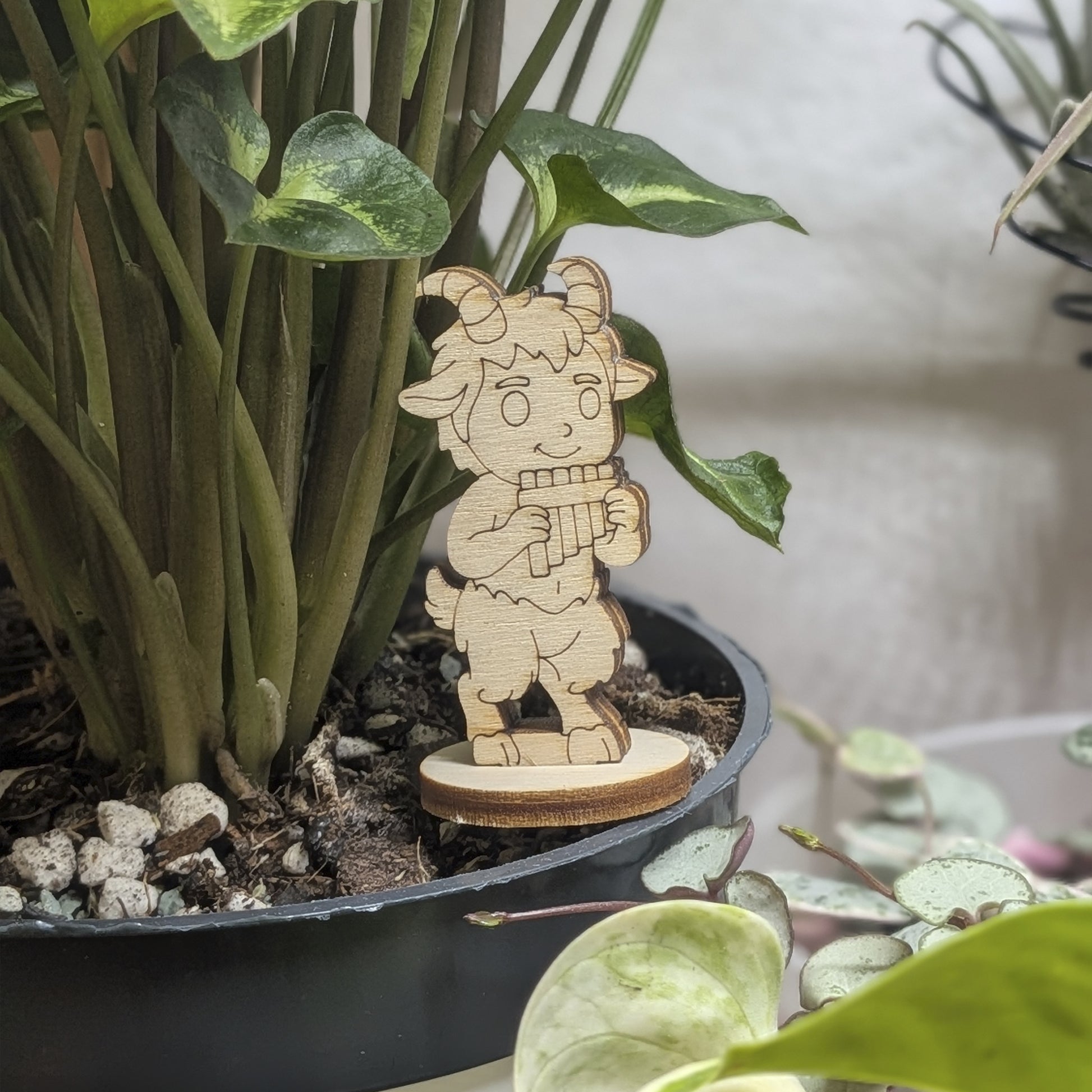 Laser cut plywood faun figurine staged in a plant pot. He plays panpipes amidst green leaves on a sturdy round wood base