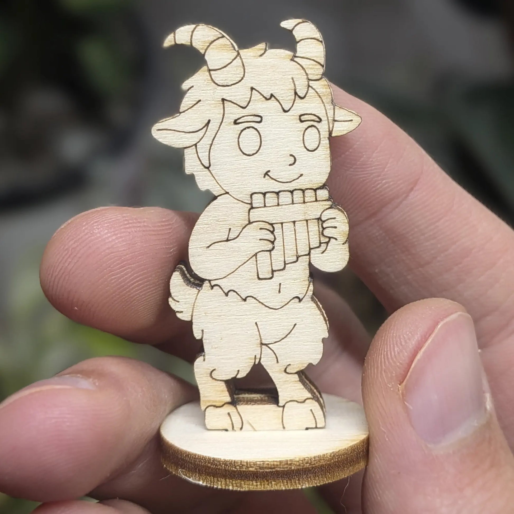 Hand holds a small faun figurine of laser cut plywood. He plays panpipes and has cute horns, furry legs, and tiny hooves