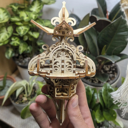 Hand holds a mini floral fairy house of laser cut plywood. Features an ornate arched door and layered scrollwork roof decor.