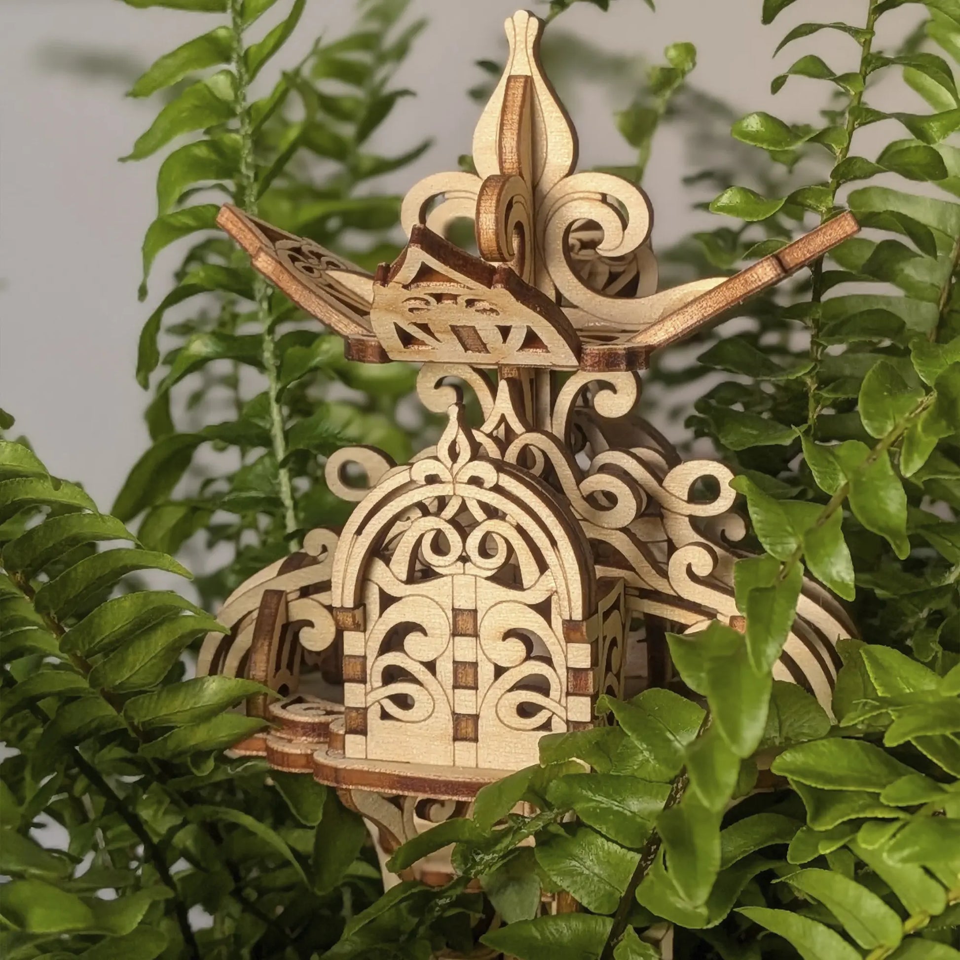 Laser cut plywood floral fairy house nestled in green fern leaves, highlighting the delicate, ornate wooden carvings here.