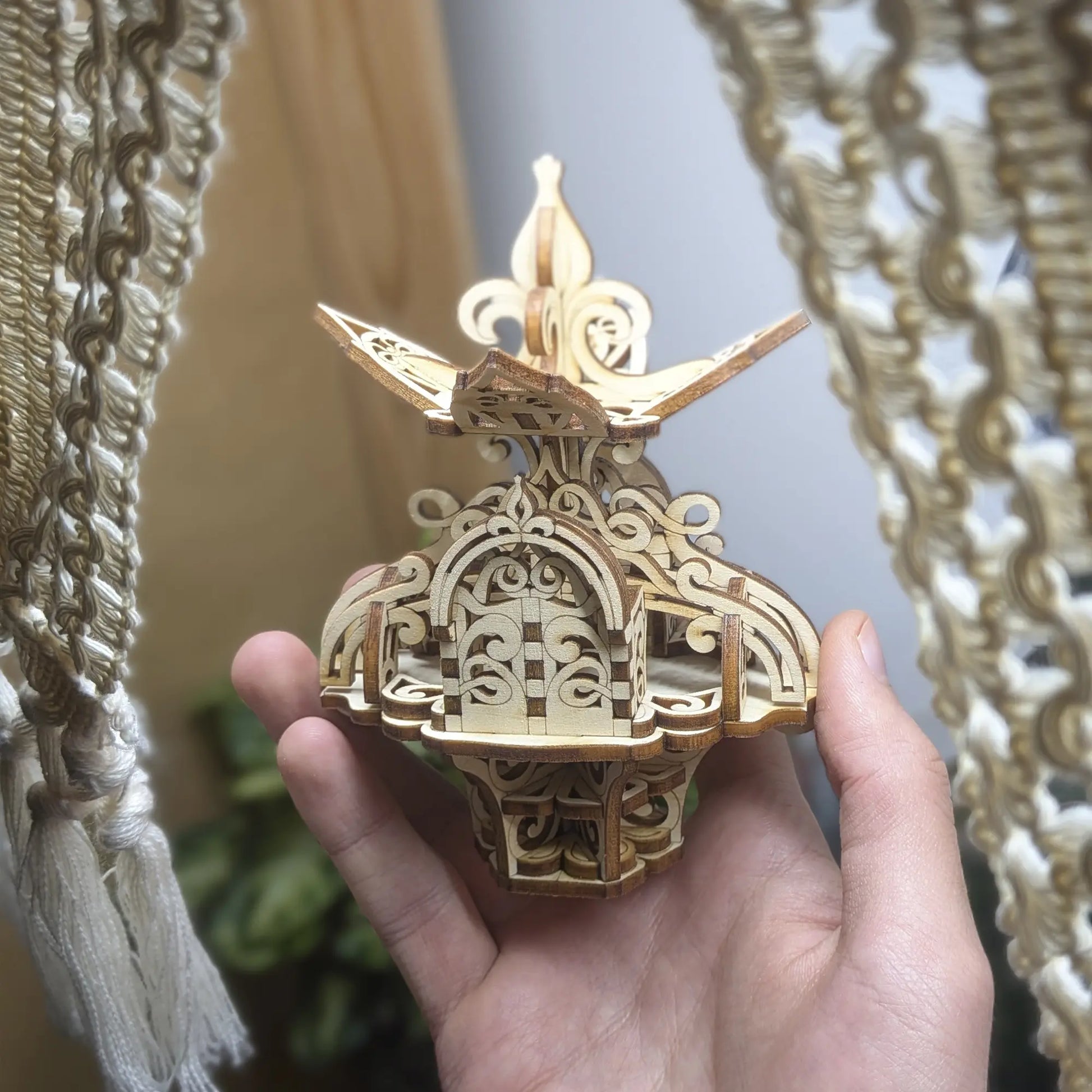 Hand holds the laser cut plywood floral fairy house near plants, showing the model's scale and intricate 3D wood structure.