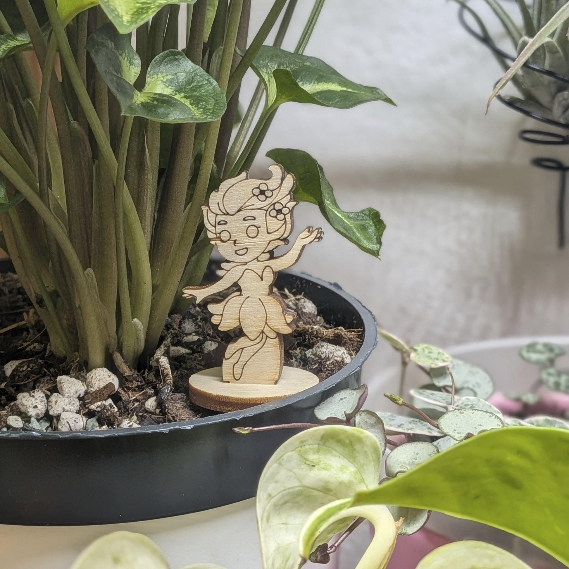 A small flower fairy girl of laser cut plywood stands in a green potted plant, featuring tiny flowers in her curly hair