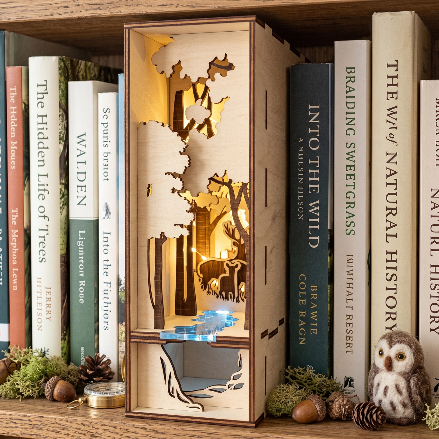 Lit laser-cut birch plywood forest book nook on a shelf with nature books, moss, pinecones, and a tiny toy owl figurine