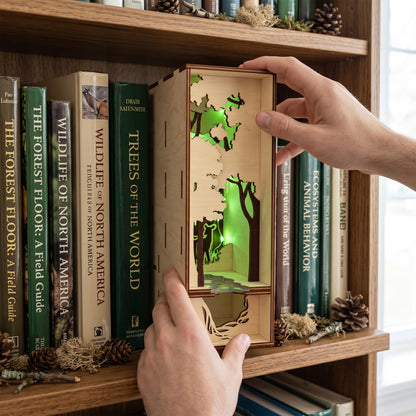 Lit laser-cut birch plywood forest book nook on a shelf between nature books, with green internal lighting and pinecones