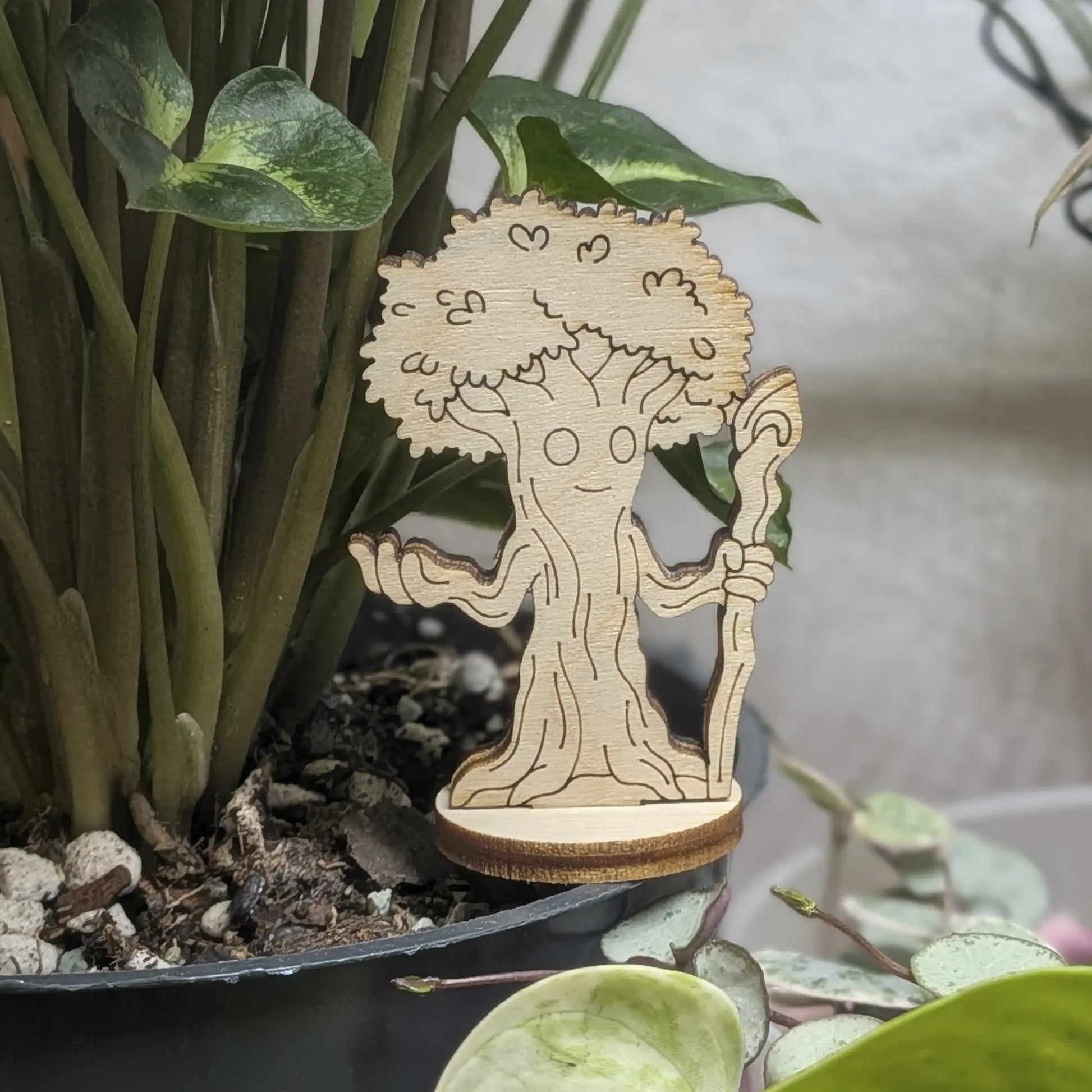 A laser cut plywood forest guardian tree figurine sits in a plant pot. Features a grumpy face, roots, and leafy branches