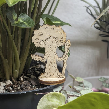 A laser cut plywood forest guardian tree figurine sits in a plant pot. Features a grumpy face, roots, and leafy branches