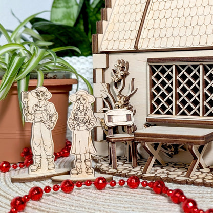 A close-up shot of two miniature wooden gardener figurines standing outside an assembled laser cut plywood garden cottage.