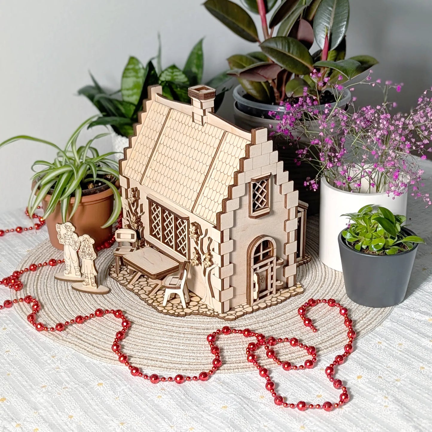 Assembled laser cut plywood garden cottage on a table with a tiny wooden patio set, tiny human figures, and house plants.